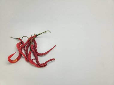 Beyaz arka planda kurumuş kırmızı biber kümesi uzanır. Biberler buruşmuş ve buruşmuş, kurumuş hallerini sergiliyorlar. Resim, genellikle yemek pişirmede kullanılan kırmızı biberlerin dokusunu ve rengini vurgular..
