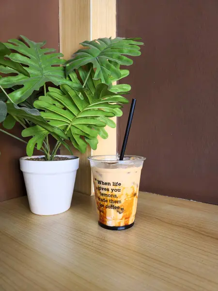 Açık plastik bir fincanda siyah pipetle buzlu kahvenin davetkar bir görüntüsü. Bardak, büyük yeşil yapraklı bir saksı bitkisinin yanındaki tahta bir yüzeye yerleştirilir. Kahvenin içinde karamel benzeri bir girdap var ve fincanda da bir alıntı var..