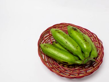 Beyaz arka planda kırmızı ve kahverengi dokuma sepetinde yeşil muzların olduğu yüksek açılı bir stüdyo çekimi. Tropikal meyveler, olgunlaşmamış muz, sağlıklı yemek ve vejetaryen diyet kavramı..