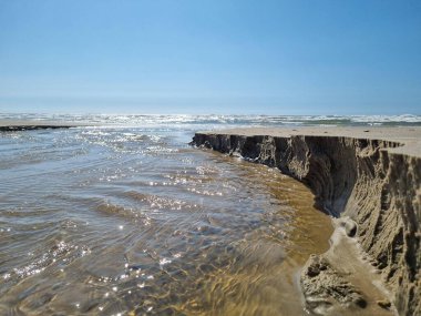 Berrak mavi bir gökyüzünün altında kumlu bir sahilin üzerindeki gel-git sularının yumuşak akışını tasvir eden sakin bir sahil manzarası. Güneş ışığı suyun yüzeyinde parlar ve kumun pürüzlü dokuları görünür..