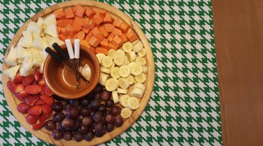 Dairesel ahşap tepside canlı bir meyve tabağı ananas, papaya, muz, çilek ve üzümler düzenlenir. Çatallı yemek çubukları ve küçük kahverengi bir kâse eşliğinde, bu ekran zarif bir yeşil ve beyaz houndstooth masa örtüsünün üzerinde duruyor. 