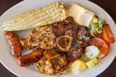 Ekvador barbeküsü, tavuk, sosis ve sığır eti karışımı, yanında mısır, taze peynir, soslu patates ve domates salatası ile birlikte beyaz bir tabakta servis edilir. Lezzetli bir And yemeği..
