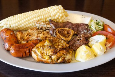 Ekvador barbeküsü, tavuk, sosis ve sığır eti karışımı, yanında mısır, taze peynir, soslu patates ve domates salatası ile birlikte beyaz bir tabakta servis edilir. Lezzetli bir And yemeği..