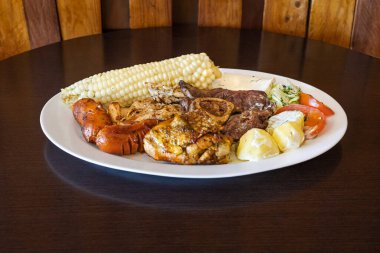 Ekvador barbeküsü, tavuk, sosis ve sığır eti karışımı, yanında mısır, taze peynir, soslu patates ve domates salatası ile birlikte beyaz bir tabakta servis edilir. Lezzetli bir And yemeği..