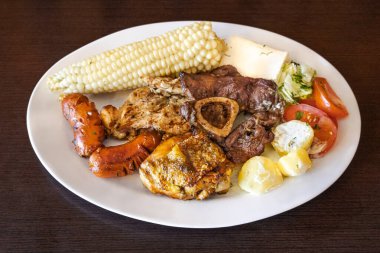 Ekvador barbeküsü, tavuk, sosis ve sığır eti karışımı, yanında mısır, taze peynir, soslu patates ve domates salatası ile birlikte beyaz bir tabakta servis edilir. Lezzetli bir And yemeği..