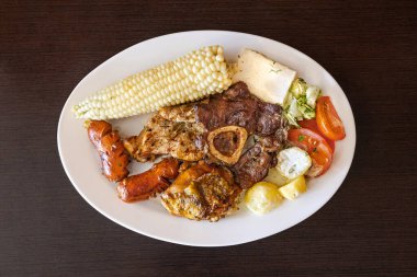 Beyaz bir tabakta servis edilen Ekvador rostosunun yanında tavuk, sosis ve sığır eti karışımı mısır, taze peynir, soslu patates ve domates salatası var. Lezzetli bir And yemeği..