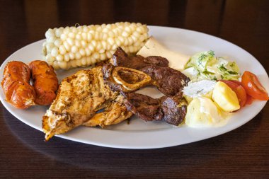 Beyaz bir tabakta servis edilen Ekvador rostosunun yanında tavuk, sosis ve sığır eti karışımı mısır, taze peynir, soslu patates ve domates salatası var. Lezzetli bir And yemeği..