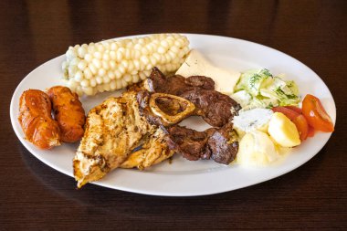 Beyaz bir tabakta servis edilen Ekvador rostosunun yanında tavuk, sosis ve sığır eti karışımı mısır, taze peynir, soslu patates ve domates salatası var. Lezzetli bir And yemeği..