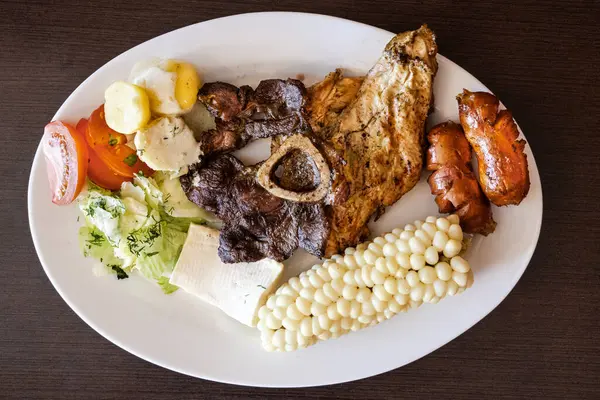 Ekvador barbeküsü, tavuk, sosis ve sığır eti karışımı, yanında mısır, taze peynir, soslu patates ve domates salatası ile birlikte beyaz bir tabakta servis edilir. Lezzetli bir And yemeği..