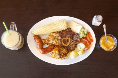 Tavuk, sığır eti ve sosisli Ekvador rostosu, mısır, taze peynir, soslu patates ve domates salatası ile servis ediliyor. Yanında naranjilla ve yulaf ezmeli colada var..