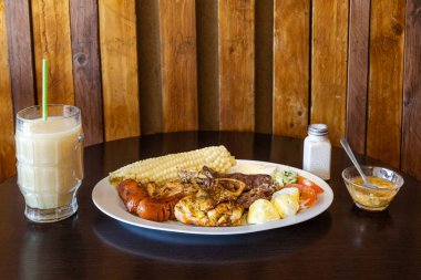 Tavuk, sığır eti ve sosisli Ekvador rostosu, mısır, taze peynir, soslu patates ve domates salatası ile servis ediliyor. Yanında naranjilla ve yulaf ezmeli colada var..