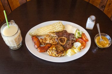 Tavuk, sığır eti ve sosisli Ekvador rostosu, mısır, taze peynir, soslu patates ve domates salatası ile servis ediliyor. Yanında naranjilla ve yulaf ezmeli colada var..