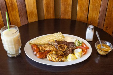Tavuk, sığır eti ve sosisli Ekvador rostosu, mısır, taze peynir, soslu patates ve domates salatası ile servis ediliyor. Yanında naranjilla ve yulaf ezmeli colada var..