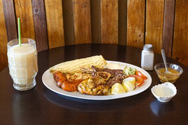 Tavuk, sığır eti ve sosisli Ekvador rostosu, mısır, taze peynir, soslu patates ve domates salatası ile servis ediliyor. Yanında naranjilla ve yulaf ezmeli colada var..