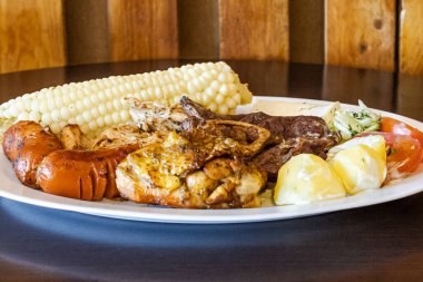 Beyaz bir tabakta servis edilen Ekvador rostosunun yanında tavuk, sosis ve sığır eti karışımı mısır, taze peynir, soslu patates ve domates salatası var. Lezzetli bir And yemeği..