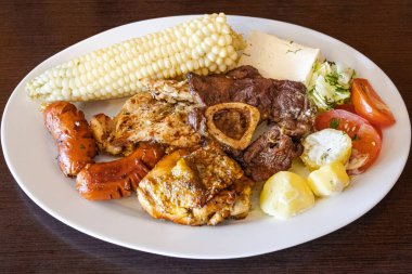 Beyaz bir tabakta servis edilen Ekvador rostosunun yanında tavuk, sosis ve sığır eti karışımı mısır, taze peynir, soslu patates ve domates salatası var. Lezzetli bir And yemeği..