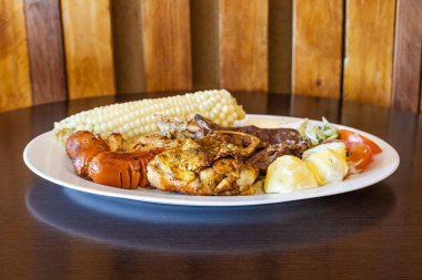 Beyaz bir tabakta servis edilen Ekvador rostosunun yanında tavuk, sosis ve sığır eti karışımı mısır, taze peynir, soslu patates ve domates salatası var. Lezzetli bir And yemeği..