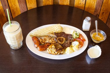 Tavuk, sığır eti ve sosisli Ekvador rostosu, mısır, taze peynir, soslu patates ve domates salatası ile servis ediliyor. Yanında naranjilla ve yulaf ezmeli colada var..