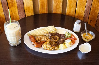 Tavuk, sığır eti ve sosisli Ekvador rostosu, mısır, taze peynir, soslu patates ve domates salatası ile servis ediliyor. Yanında naranjilla ve yulaf ezmeli colada var..