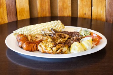 Beyaz bir tabakta servis edilen Ekvador rostosunun yanında tavuk, sosis ve sığır eti karışımı mısır, taze peynir, soslu patates ve domates salatası var. Lezzetli bir And yemeği..