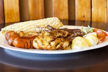 Beyaz bir tabakta servis edilen Ekvador rostosunun yanında tavuk, sosis ve sığır eti karışımı mısır, taze peynir, soslu patates ve domates salatası var. Lezzetli bir And yemeği..