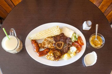 Tavuk, sığır eti ve sosisli Ekvador rostosu, mısır, taze peynir, soslu patates ve domates salatası ile servis ediliyor. Yanında naranjilla ve yulaf ezmeli colada var..