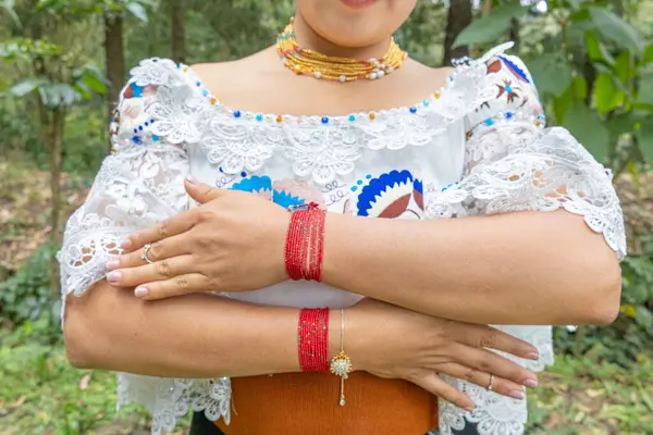 Otavalea yerlisi bir kadın kırmızı bileziğini ya da manillasını gösteriyor, anakolu geleneksel kıyafetler giyiyor, nakışlı bluz, altın walka kolyesi, kahverengi manila kuşağı.