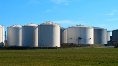 Several large storage tanks stand side by side at an industrial facility. They are surrounded by lush green fields under a bright blue sky, showcasing a clear midday atmosphere.