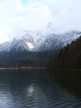 Karla kaplı dağlar sakin bir göl üzerinde görkemli bir şekilde yükselir. Sahne sakin, suda hafif dalgalanmalar ve serin, bulutlu bir atmosfer var..