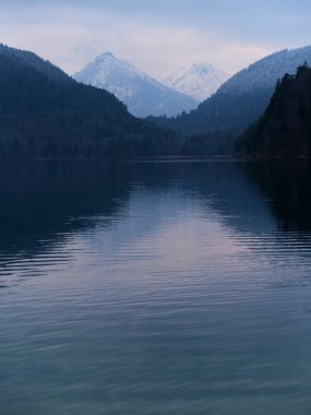 Gölün sakin suları, alacakaranlıkta yemyeşil ve dağlarla çevrili uzaktaki karlı tepeleri yansıtıyor. Huzurlu bir atmosfer yaratıyor..