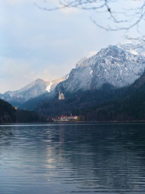 Sakin bir göl, karla kaplı dağları ve uzak bir yapıyı yansıtır. Kış boyunca huzur ve doğal güzelliği çağrıştırır..