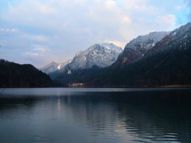 Sakin bir kış manzarası çevresindeki dağları yansıtan sakin bir göle sahiptir. Arka planda karla kaplı zirveler yükselirken, uzak bir bina manzaraya ilgi gösteriyor.