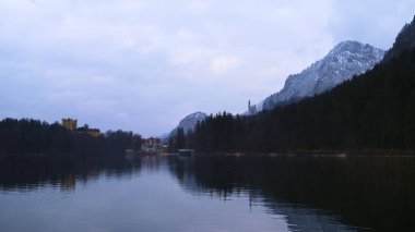 Sakin sular, karlı dağları ve alacakaranlıkta büyüleyici bir köyü yansıtır. Çam ağaçları kıyı boyunca uzanır, huzurlu bir kış ortamı yaratır, sükuneti ve yansımayı davet eder..