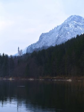 Durgun bir göl üzerinde yükselen dağ tepelerini kar örtüyor. Uzakta bir kale beliriyor, sakin bir gökyüzünün altında sık ve yeşil ormanlarla çevrili..