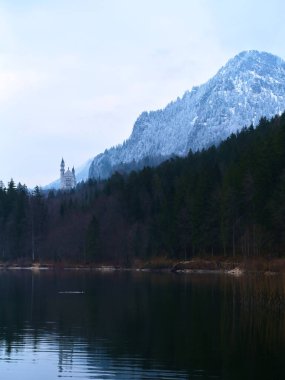 Kar ormanı ve sakin bir gölü çevreleyen dağların tepelerini kaplıyor. Neuschwanstein Şatosu 'nun uzak kuleleri kış manzarasından ortaya çıkarak pitoresk bir manzara yaratıyor..
