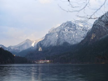 Karla kaplı dağlar, güzelliklerini yansıtan sakin bir göl üzerinde belirir. Kıyı boyunca uzanan büyüleyici bir kale, alacakaranlıktaki bu manzaranın sükunetini artırır..