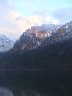 Karla kaplı dağlar sakin suların üzerinde yükselir, yumuşak sabah ışığını yansıtır. Bulutlar tembelce gökyüzünde sürüklenirken doğa huzurlu güzelliğini sergiliyor, tefekküre ve keşfe davet ediyor..