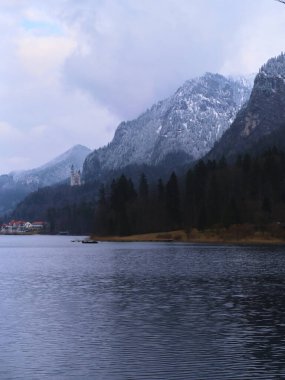 Huzurlu bir kış sabahı, karla kaplı dağlarla çevrili sessiz bir göl ve kıyı şeridi boyunca küçük bir köy sergileniyor. Yumuşak bulutlar durgun manzaranın üzerinde duruyor..