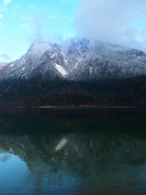 Karla kaplı dağlar mavi gökyüzüne karşı dimdik dururlar. Yansımaları aşağıdaki sakin gölde açıkça görülür. Sabah ışığı doğaya gölge düşürür..