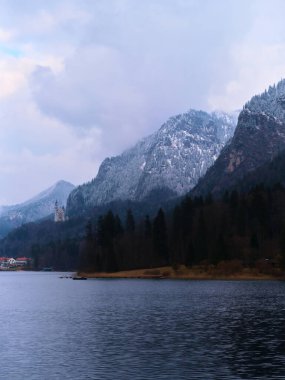 Huzurlu bir kış manzarası, sakin bir göl üzerinde karla kaplı dağların yükseldiğini gösteriyor. Çamlar kıyı boyunca dimdik durur, yansıma için mükemmel sakin bir atmosfer yaratır..