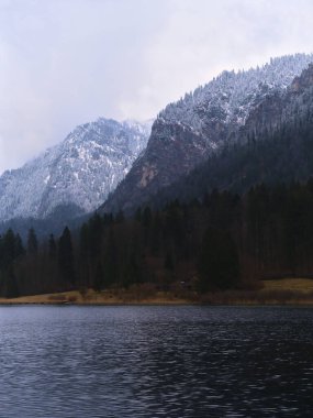 Karla kaplı zirveler sakin bir göl üzerinde görkemli bir şekilde yükselir ve huzurlu bir kış ortamı yaratır. Durgun su bulutlu bir gökyüzünün altındaki buzlu dağları yansıtır..