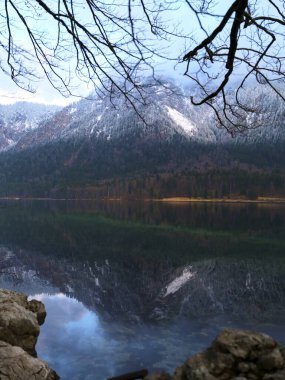 Gölün sakin suları görkemli karla kaplı dağları ve çıplak ağaç dallarını yansıtıyor. Sabahın erken saatlerinde yumuşak bir ışık uzak bir doğal ortamda sakin bir atmosfer yaratır..