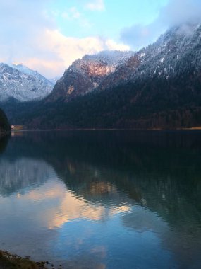 Durgun bir göl, sabahın erken saatlerinde renkli bir gökyüzü altında karla kaplı dağların yansımasını yakalar. Huzurlu ortam kış doğasının güzelliğini gözler önüne seriyor..