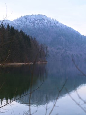 Sabahın erken saatlerinde, karla kaplı bir dağ sakin bir gölün üzerinde görkemli bir şekilde duruyor, yansıması su yüzeyinde parıldıyor. Ağaçlar gölün kenarını çiziyor, sakin manzarayı güçlendiriyor..