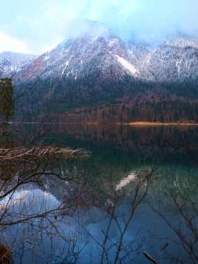 Karla kaplı dağlarla çevrili sakin bir gölü gösteren huzurlu bir manzara. Sabahın erken saatlerinde yansıtıcı suya yumuşak bir parıltı yayar, dingin atmosferi yükseltir..