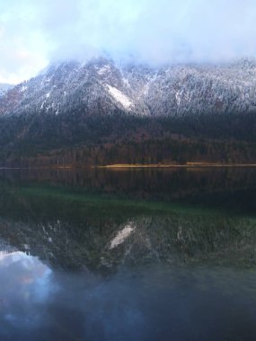 Karla kaplı dağlar sakin bir göl üzerinde yükselir, yansımaları su üzerinde aynaya benzer bir etki yaratır. Bulutlar gökyüzünde süzülüyor, sakin atmosferi güçlendiriyor..