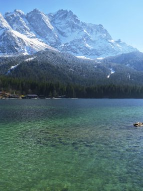 Görkemli dağlar yeşil ormanlarla çevrili sakin bir turkuaz göl üzerinde kule gibi. Sahne, parlak güneş ışığı ve canlı renklerle dolu sakin bir yaz gününü yakalıyor..