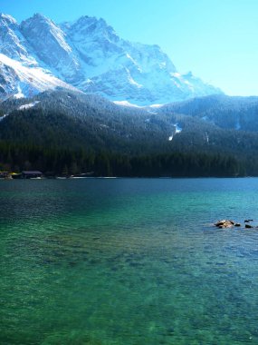 Görkemli dağlar sakin bir göl üzerinde kule gibi, karla kaplı zirveleri güneş ışığında parıldıyor. Kristal berrak sular, sahil boyunca kayalara nazikçe çarpar ve huzurlu bir kaçış sunar..