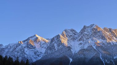 Dağlar görkemli bir şekilde yükselir, taze karla kaplı, el değmemiş mavi gökyüzünün altında. Güneşin altın renkleri tepeleri aydınlatıyor, nefes kesici bir dağ manzarası yaratıyor..