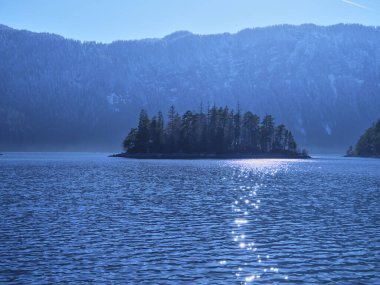 Açık mavi suyla çevrili küçük bir adanın huzurlu görüntüsü güneş ışığını yansıtır. Görkemli dağlar parlak bir gökyüzünün altında arka planda yükseliyor ve sakin bir atmosfer yaratıyor..